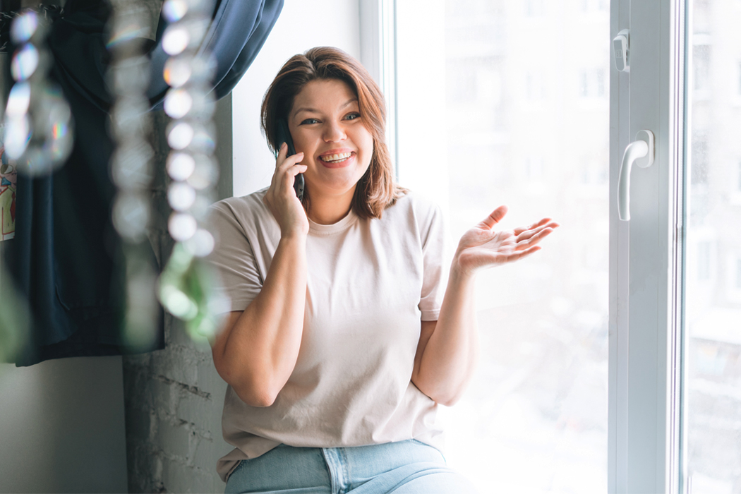 A smiling woman talking on the phone in front of a brightly lit window